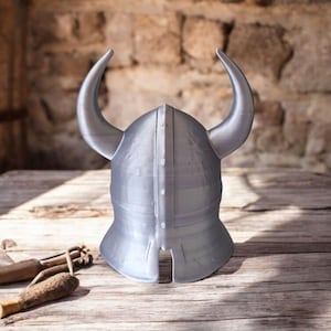 May include: A silver-colored Viking helmet with curved horns, resting on a wooden surface. The helmet is the focal point, showcasing its design and craftsmanship. The background features a rustic stone wall, enhancing the historical theme.