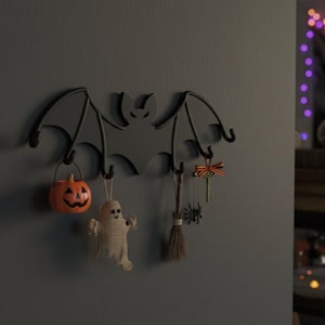 May include: A black metal bat-shaped wall hook with four hooks, decorated with Halloween-themed ornaments. The ornaments include a small orange pumpkin bucket, a burlap ghost, a broom, a key, and a spider.