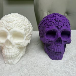 May include: Two decorative skull figurines. One is white with floral patterns, the other is purple with similar detailing. The skulls are placed side by side on a light-colored surface, showcasing their intricate designs.