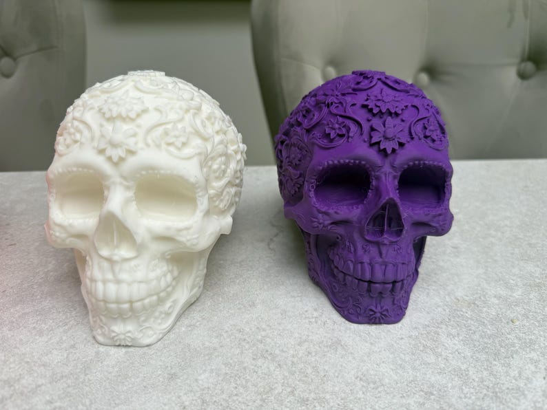 May include: Two decorative skull figurines. One is white with floral patterns, the other is purple with similar detailing. The skulls are placed side by side on a light-colored surface, showcasing their intricate designs.