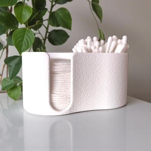 May include: A white, textured, rectangular container with rounded edges, designed to hold cotton pads and cotton swabs. The container is divided into two sections, one for cotton pads and the other for cotton swabs. A green plant is in the background.