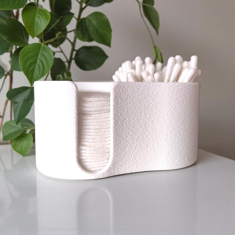 May include: A white, textured, rectangular container with rounded edges, designed to hold cotton pads and cotton swabs. The container is divided into two sections, one for cotton pads and the other for cotton swabs. A green plant is in the background.