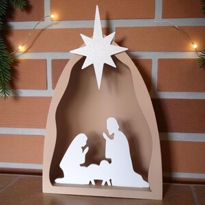 May include: A wooden nativity scene with a star on top. The scene features silhouettes of Mary, Joseph, and baby Jesus inside a beige arch. The star is white and glittery. The background is a brick wall.