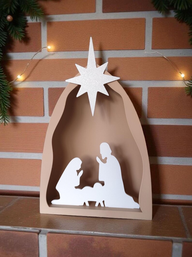 May include: A wooden nativity scene with a star on top. The scene features silhouettes of Mary, Joseph, and baby Jesus inside a beige arch. The star is white and glittery. The background is a brick wall.