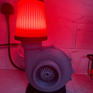 May include: A unique lamp with a red glowing lampshade and a gray turbine engine base. The lamp has a black base and a power cord. The turbine has a fan-like design in the center.