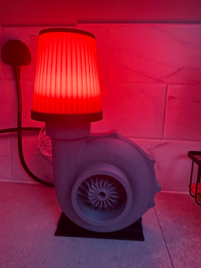 May include: A unique lamp with a red glowing lampshade and a gray turbine engine base. The lamp has a black base and a power cord. The turbine has a fan-like design in the center.