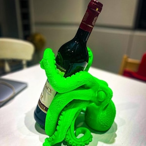 May include: A bright green octopus-shaped wine bottle holder. The tentacles wrap around a dark wine bottle with a red and gold label. The holder is on a white surface, creating a striking contrast.