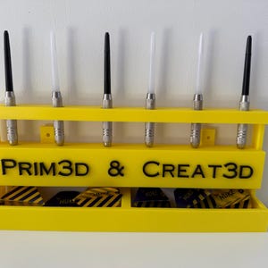 Puede incluir: Un soporte de dardos amarillo brillante con seis dardos y un compartimento inferior para plumas. El soporte tiene el texto "PRIM3D & CREATE3D" en negro. Los dardos tienen barriles de metal plateado y ca&ntilde;as negras o blancas.