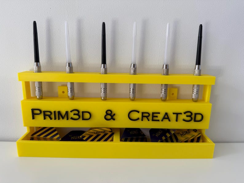 Puede incluir: Un soporte de dardos amarillo brillante con seis dardos y un compartimento inferior para plumas. El soporte tiene el texto "PRIM3D & CREATE3D" en negro. Los dardos tienen barriles de metal plateado y ca&ntilde;as negras o blancas.