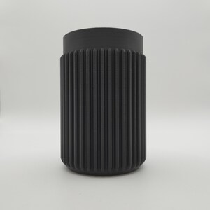 May include: A cylindrical black container with a ribbed exterior. The container has a slightly wider top and a rounded base. The vertical ribs create a textured surface. The object is set against a plain white background.