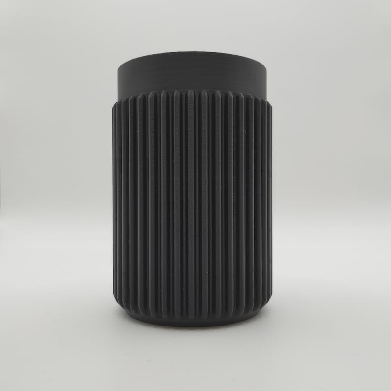 May include: A cylindrical black container with a ribbed exterior. The container has a slightly wider top and a rounded base. The vertical ribs create a textured surface. The object is set against a plain white background.