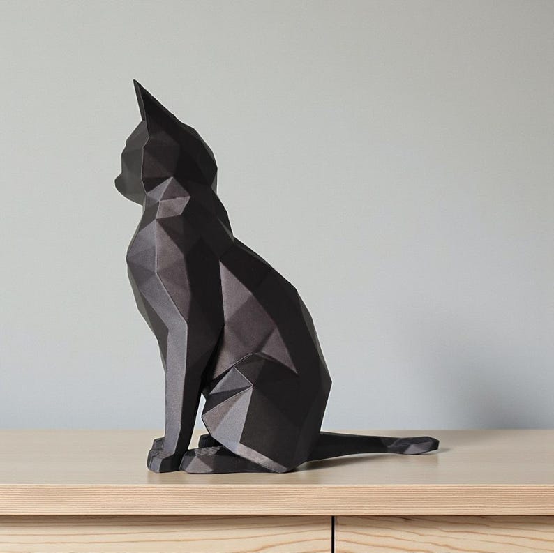 May include: A black geometric cat sculpture, sitting upright with its tail extended. The cat is made of angular, flat surfaces, creating a modern, polygonal design. The sculpture is placed on a light-colored wooden surface.
