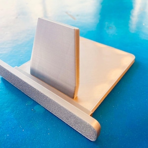 May include: A silver-coloured phone stand with a textured base and a vertical support. The stand has a rectangular base and a back support that is angled slightly backward. The stand is made of metal and sits on a blue surface.