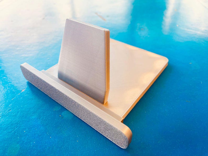 May include: A silver-coloured phone stand with a textured base and a vertical support. The stand has a rectangular base and a back support that is angled slightly backward. The stand is made of metal and sits on a blue surface.