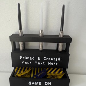 May include: A black dart holder with three darts and a compartment for dart flights. The holder has the text "Prim3d & Creat3d Your Text Here" and "GAME ON". The dart flights are yellow and blue.