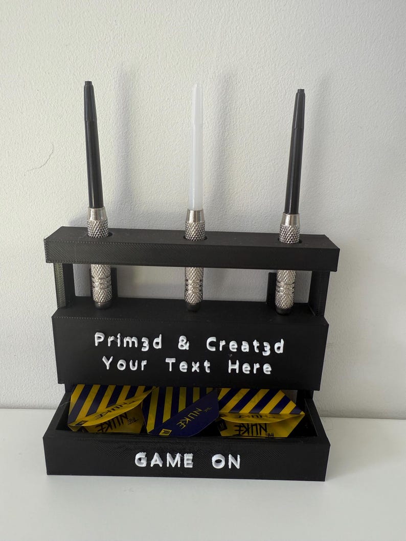 May include: A black dart holder with three darts and a compartment for dart flights. The holder has the text "Prim3d & Creat3d Your Text Here" and "GAME ON". The dart flights are yellow and blue.