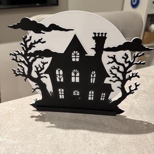 Pu&ograve; includere: Decorazione di Halloween in bianco e nero raffigurante la silhouette di una casa infestata con la luna piena. La casa ha pi&ugrave; finestre e un camino, incorniciata da alberi spogli e nuvole, creando una scena spettrale.
