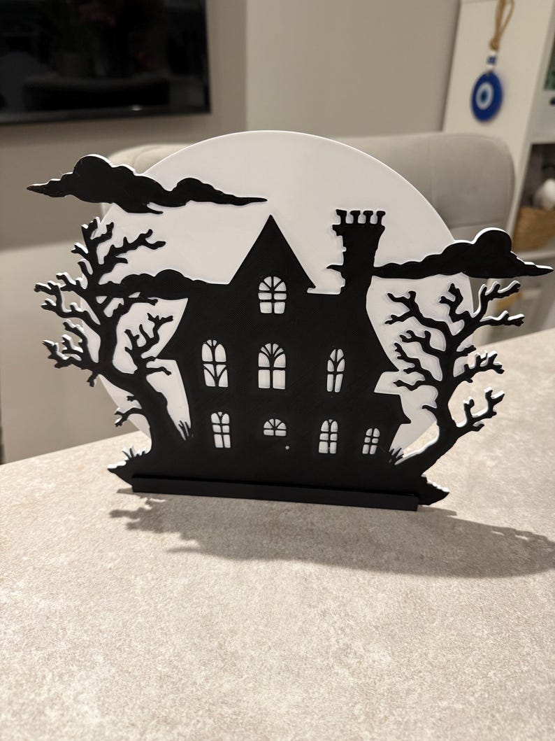 Pu&ograve; includere: Decorazione di Halloween in bianco e nero raffigurante la silhouette di una casa infestata con la luna piena. La casa ha pi&ugrave; finestre e un camino, incorniciata da alberi spogli e nuvole, creando una scena spettrale.