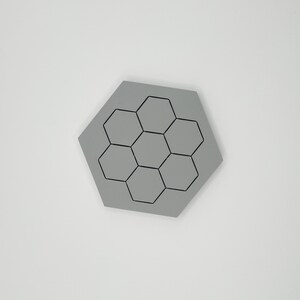 May include: A grey, hexagonal coaster with a honeycomb design. The coaster features a series of black outlines of hexagons arranged in a central pattern. The coaster is a solid grey colour.