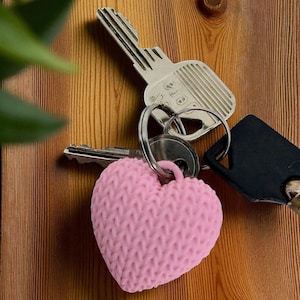 May include: A collection of keys, including a pink heart-shaped keyring with a knitted texture. The keys are silver and black, and are arranged on a wooden surface. The image is a close-up shot.