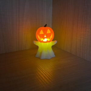 May include: A glowing orange jack-o'-lantern with a carved face sits atop a white, ghost-shaped base. The Halloween decoration emits a warm, yellow light, creating a festive ambiance. The pumpkin has a black stem.