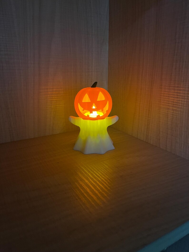 May include: A glowing orange jack-o'-lantern with a carved face sits atop a white, ghost-shaped base. The Halloween decoration emits a warm, yellow light, creating a festive ambiance. The pumpkin has a black stem.
