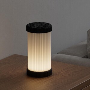 Pode incluir: Uma l&acirc;mpada cil&iacute;ndrica com parte superior e base pretas, e uma tela branca com nervuras verticais. A l&acirc;mpada est&aacute; iluminada, emitindo um brilho suave. Est&aacute; sobre uma superf&iacute;cie de madeira.