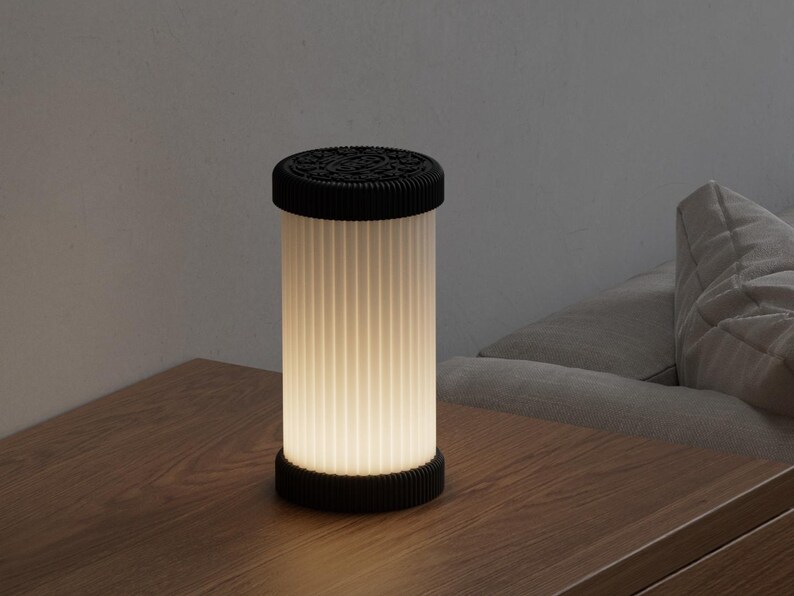Pode incluir: Uma l&acirc;mpada cil&iacute;ndrica com parte superior e base pretas, e uma tela branca com nervuras verticais. A l&acirc;mpada est&aacute; iluminada, emitindo um brilho suave. Est&aacute; sobre uma superf&iacute;cie de madeira.