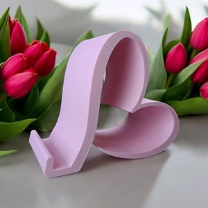 May include: A lavender heart-shaped phone stand. The stand has a cutout for a phone and a raised base. The stand is sitting on a white surface.