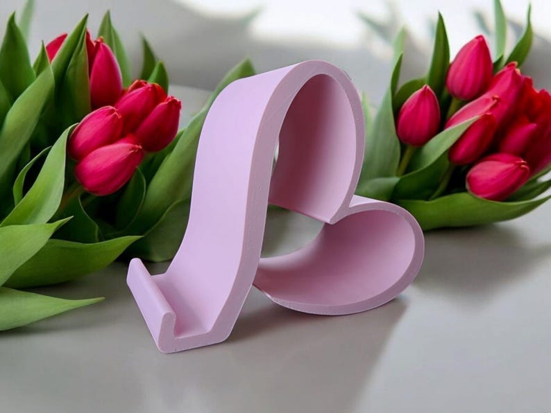 May include: A lavender heart-shaped phone stand. The stand has a cutout for a phone and a raised base. The stand is sitting on a white surface.