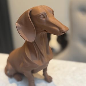 May include: A brown dachshund dog figurine, sculpted with detailed features. The dog is sitting upright, with its long body and floppy ears. The figurine is made of a solid material and has a smooth finish. The dog's eyes and nose are clearly defined.