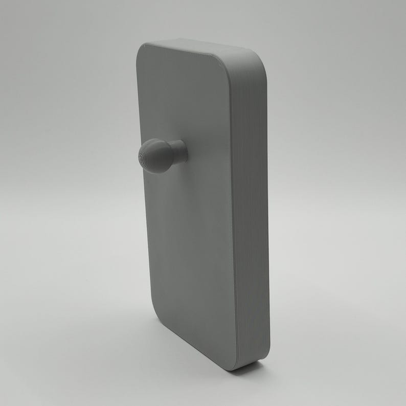K&ouml;nnte beinhalten: Ein graues, rechteckiges Objekt mit abgerundeten Ecken und einem kleinen, strukturierten Knopf, der von einer Seite herausragt. Das Objekt ist ein solides, monochromes Design, wahrscheinlich ein T&uuml;rgriff oder eine &auml;hnliche Hardware.