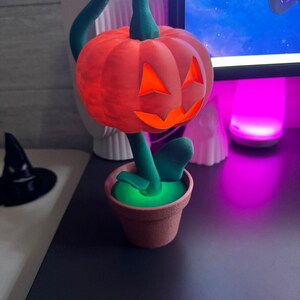 May include: A Halloween-themed decoration featuring a carved orange pumpkin with glowing orange eyes and mouth, atop a green stem and leaves. The pumpkin sits in a small, brown flower pot, creating a festive, whimsical display.