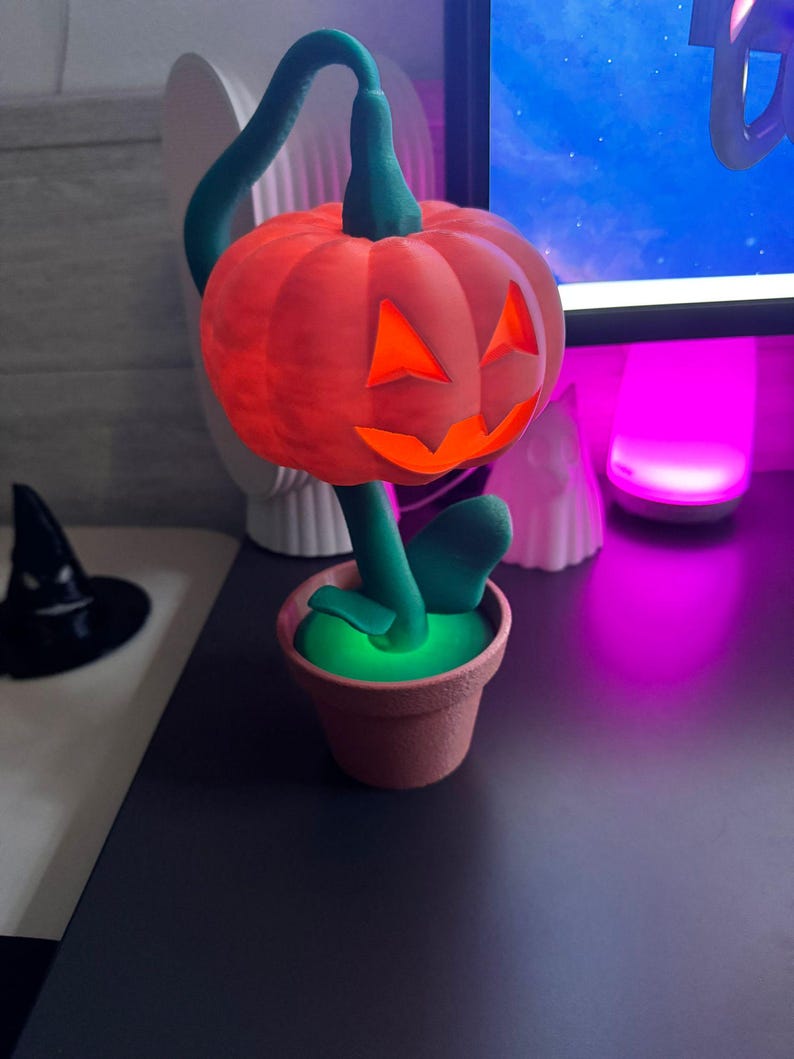 May include: A Halloween-themed decoration featuring a carved orange pumpkin with glowing orange eyes and mouth, atop a green stem and leaves. The pumpkin sits in a small, brown flower pot, creating a festive, whimsical display.
