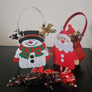 May include: Christmas-themed gift baskets shaped like a snowman and Santa Claus, with handles. The snowman has a black hat and a red and green scarf. The Santa basket is red with a white beard. Chocolate candies are in front.