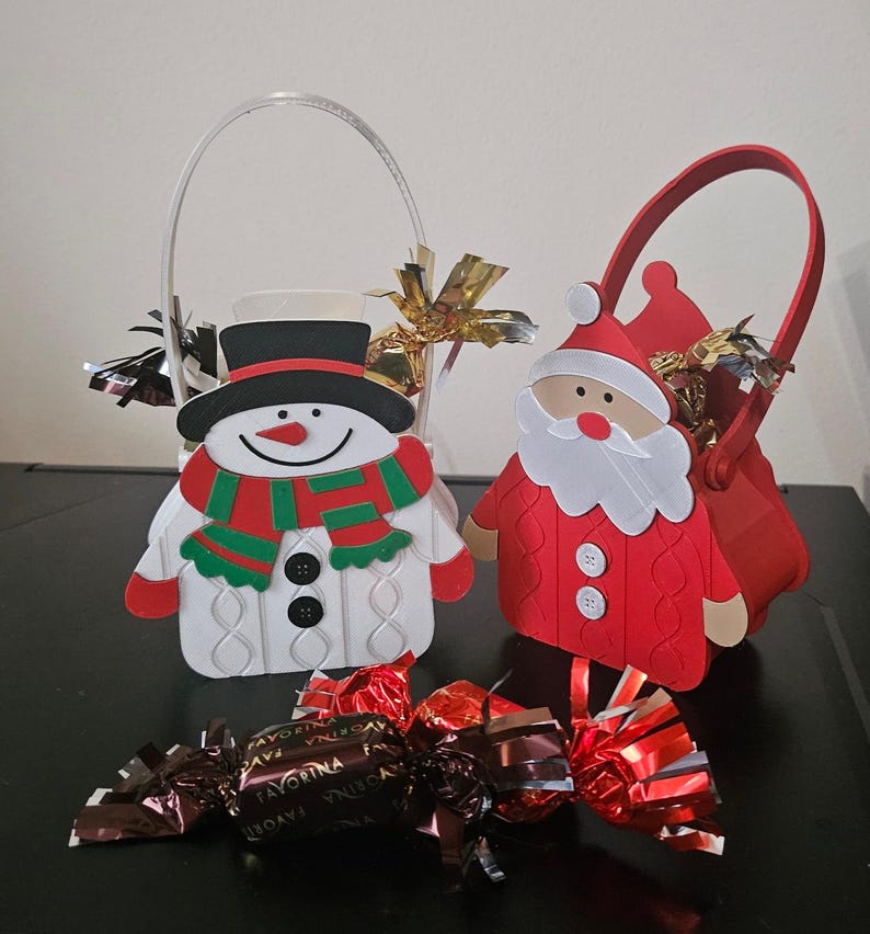 May include: Christmas-themed gift baskets shaped like a snowman and Santa Claus, with handles. The snowman has a black hat and a red and green scarf. The Santa basket is red with a white beard. Chocolate candies are in front.