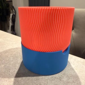 May include: A two-tone, 3D-printed planter. The upper section is a vibrant orange with a textured, wavy pattern. The base is a solid blue. The planter is designed to hold plants and add a modern touch to any space.