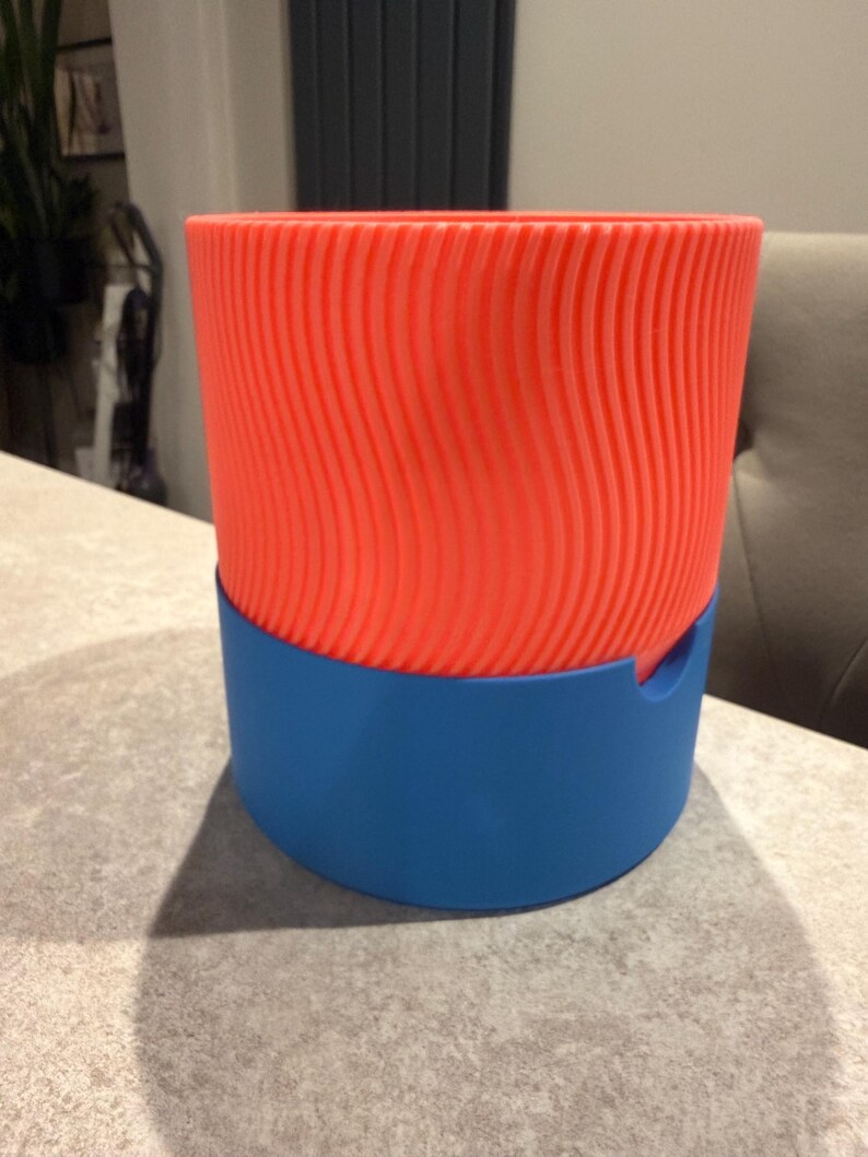 May include: A two-tone, 3D-printed planter. The upper section is a vibrant orange with a textured, wavy pattern. The base is a solid blue. The planter is designed to hold plants and add a modern touch to any space.