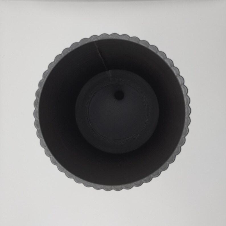May include: A black, cylindrical planter with a scalloped rim. Inside, a smaller, round, black insert with a central hole is visible. The planter is set against a plain white background, creating a stark contrast.