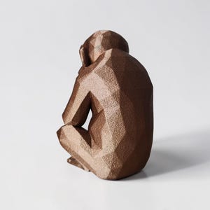 May include: A bronze-colored geometric sculpture of a seated monkey, with its head resting on its knees. The sculpture has a faceted design, giving it a modern, angular appearance. The monkey is positioned against a plain white background.
