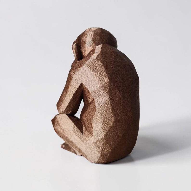 May include: A bronze-colored geometric sculpture of a seated monkey, with its head resting on its knees. The sculpture has a faceted design, giving it a modern, angular appearance. The monkey is positioned against a plain white background.
