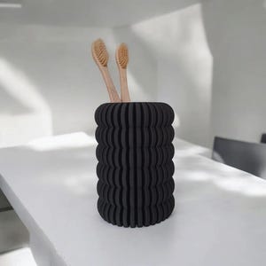 May include: A black, cylindrical toothbrush holder with a ribbed design. Two wooden toothbrushes are placed inside. The holder is on a white surface, suggesting a bathroom setting.
