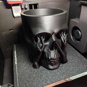 May include: A black skull-shaped planter with a wide opening and detailed facial features. The eye sockets have intricate, weeping-like designs. The planter is made of a matte black material and sits on a textured surface.