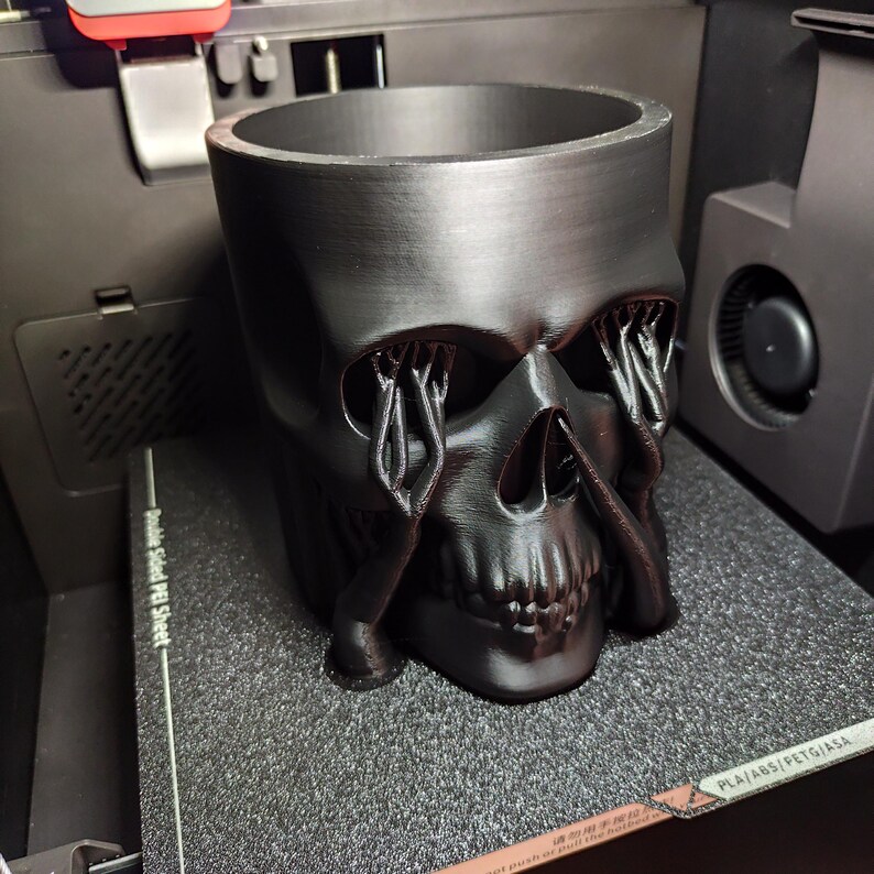 May include: A black skull-shaped planter with a wide opening and detailed facial features. The eye sockets have intricate, weeping-like designs. The planter is made of a matte black material and sits on a textured surface.