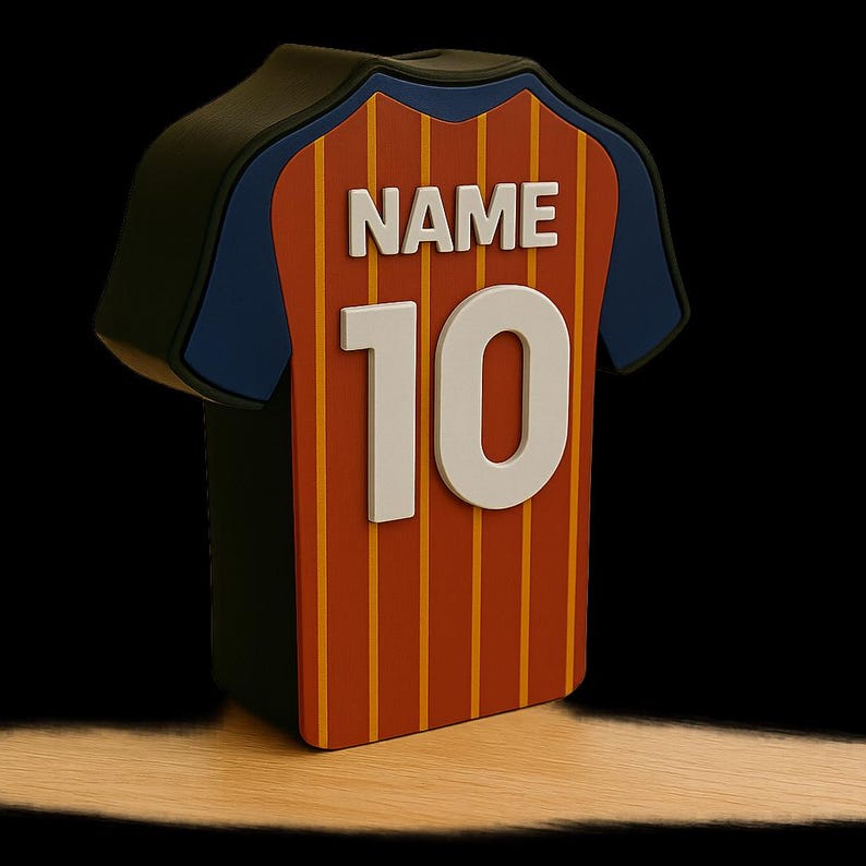 K&ouml;nnte beinhalten: Ein dekorativer Gegenstand in Trikotform mit einem rot-orangen Streifenmuster. Das Trikot hat einen blauen Kragen und &Auml;rmel, mit dem Wort "NAME" und der Nummer "10" in Wei&szlig;. Der Hintergrund ist schwarz.