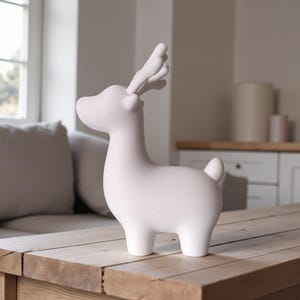 May include: A white ceramic deer figurine with detailed antlers and a small tail. The deer is standing on a wooden table. The figurine has a smooth, matte finish and is a decorative item.