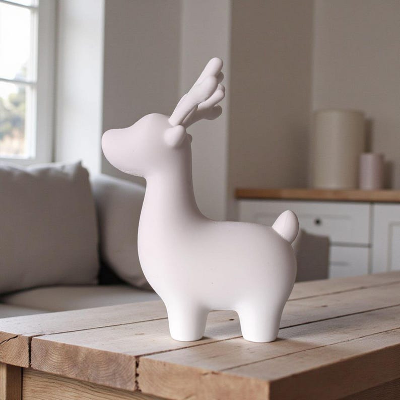 May include: A white ceramic deer figurine with detailed antlers and a small tail. The deer is standing on a wooden table. The figurine has a smooth, matte finish and is a decorative item.