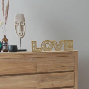 May include: A wooden dresser with the word "LOVE" in gold, rattan-style letters. Decorative items include a white mask sculpture, vases, and a black bowl. The scene is set against a neutral wall.
