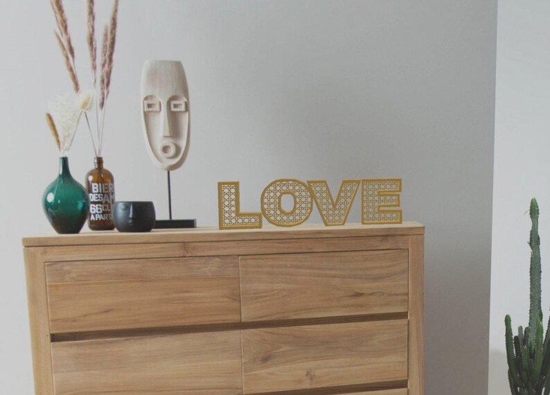 May include: A wooden dresser with the word "LOVE" in gold, rattan-style letters. Decorative items include a white mask sculpture, vases, and a black bowl. The scene is set against a neutral wall.