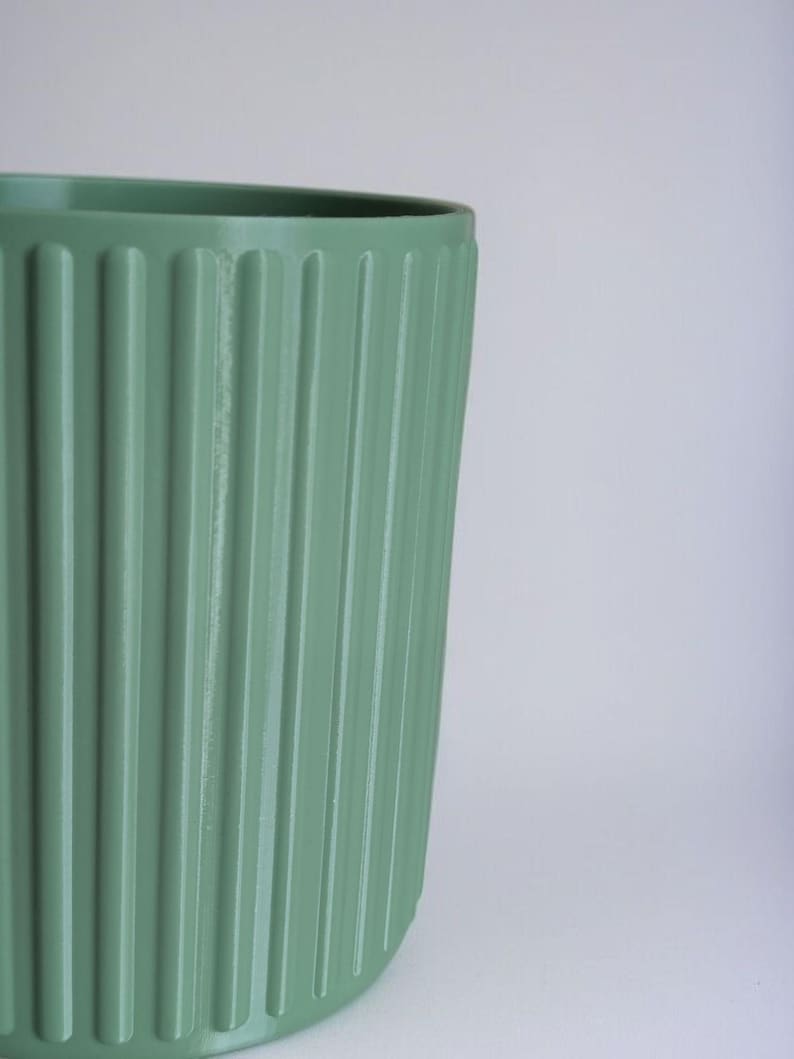 May include: A sage green ceramic planter with vertical ribbed detailing. The cylindrical pot has a rounded base and a smooth rim. The planter is designed for indoor or outdoor use and is suitable for various plants.